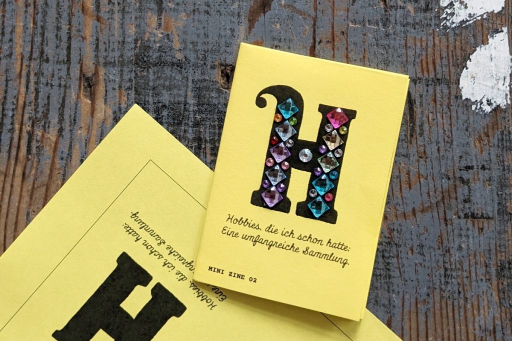 Mini Zine auf knalligem gelbem Papier. Auf dem Titel ist ein großes schwarzes gezeichnetes H, das mit bunten Edelstein-Stickern beklebt ist.
