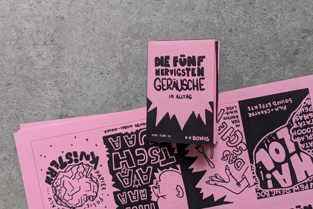 Gefaltetes Mini Zine liegt auf einem Stapel ungefalteter, rosa Papierbögen, die mit schwarzen Illustrationen bedruckt sind.
