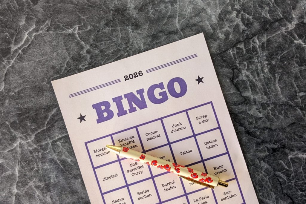 Blatt mit großem lila BINGO Schriftzug mit Sternchen und kleinen beschrifteten Quadraten liegt mit einem Stift auf einem schwarz-marmoriertem Untergrund