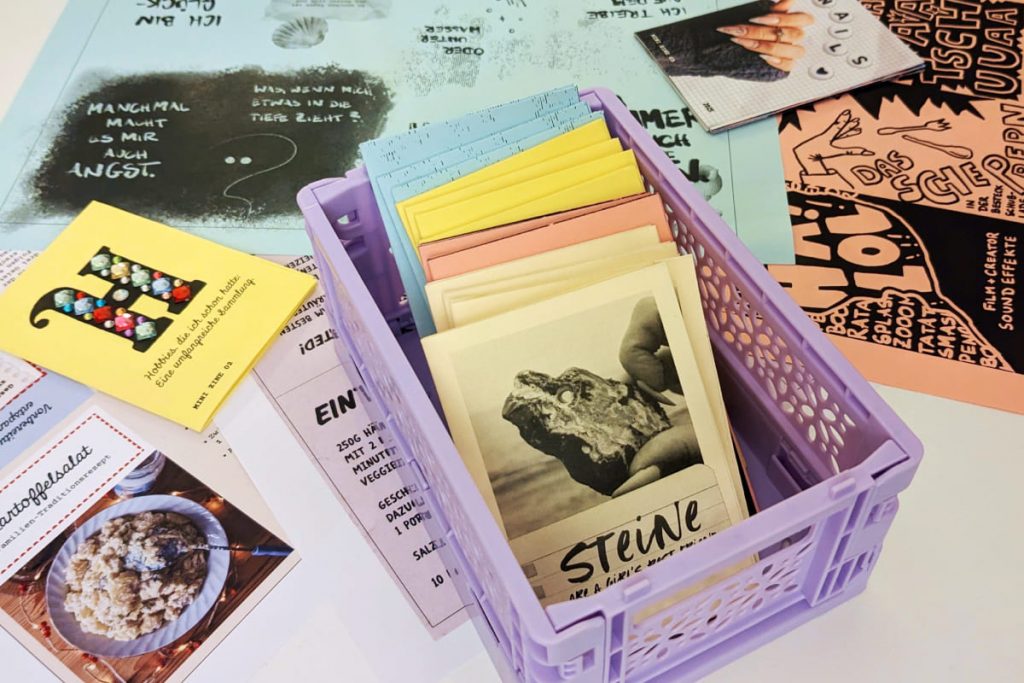 Mini-Klappkiste mit vielen kleinen Zines in bunten Farben. Rundherum liegen Blätter mit ungefalteten Zines.