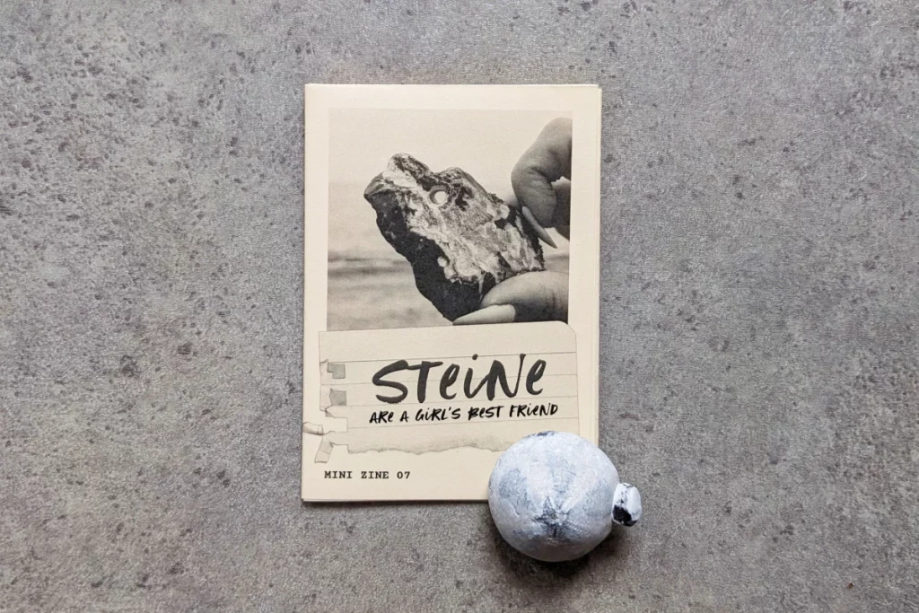 Mini Zine, auf dem versteinerter Seeigel liegt. Auf dem Cover hält eine Hand einen Stein mit Loch in den Himmel.