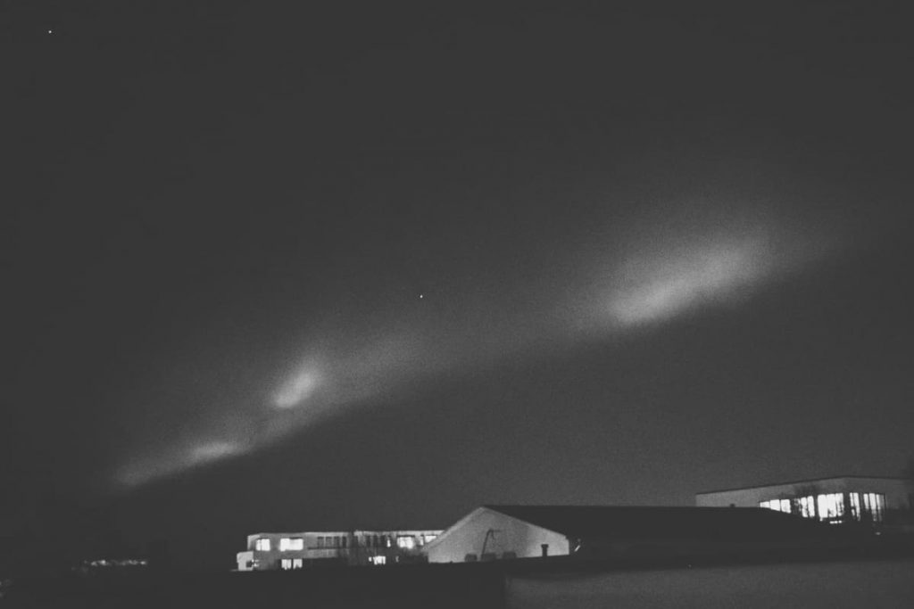 Schwarzweiß Foto von Nordlichtern, die in einem diagonalen Streifen quer über dem Himmel leuchten. Darunter sind in der Ferne Häuser zu sehen.