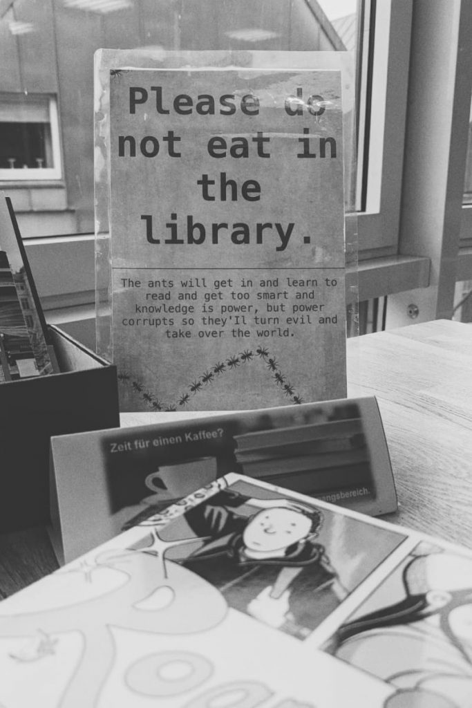 Schwarzweiß Foto, Fensterplatz, unscharfes Buch im Vordergrund. Dahinter der Fokus auf ein laminiertes Schild "Please do not eat in the library. The ants will get in and learn to read and get too smart (...)".
