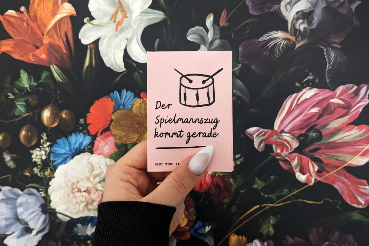Hand hält hellrotes Mini Zine. Titel mit Handschrift "Der Spielmannszug kommt gerade". Kindlich illustrierte Trommel. Im Hintergrund eine Blumentapete.