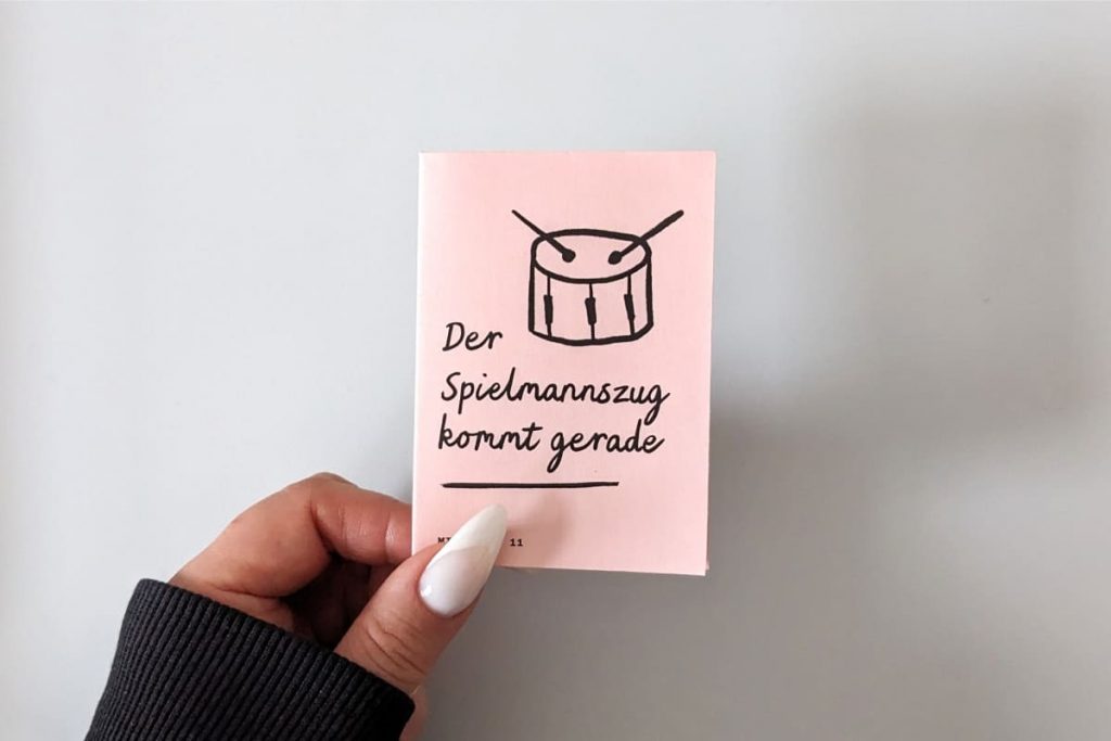 Hand hält hellrotes Mini Zine. Titel mit Handschrift "Der Spielmannszug kommt gerade". Kindlich illustrierte Trommel.
