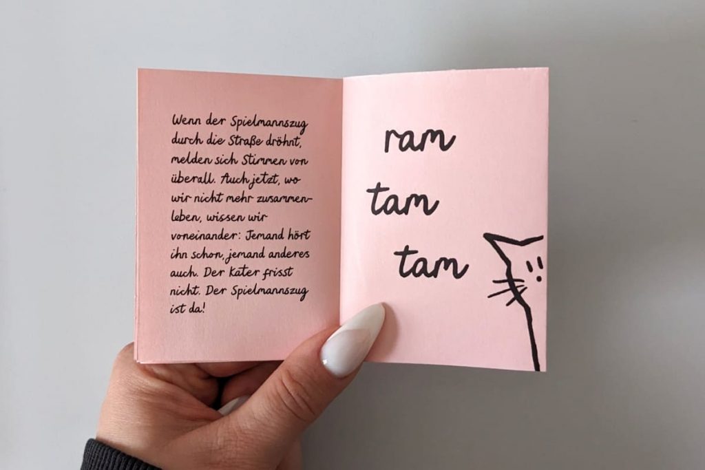 Hand hält geöffnetes Zine. Kindlich illustrierte Katze guckt skeptisch seitlich vom Rand herein. RAM TAM TAM. Alle Texte im Beitrag.