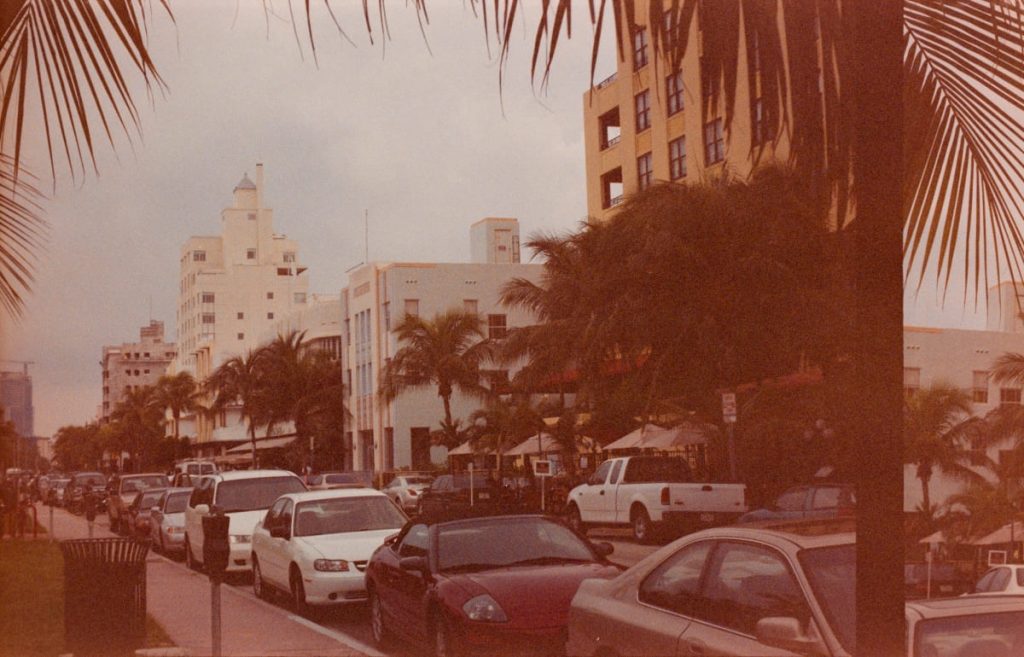 Straßenszene in Miami Beach mit Art-Déco-Häusern, Palmen und Autos bei sonnigem Wetter.