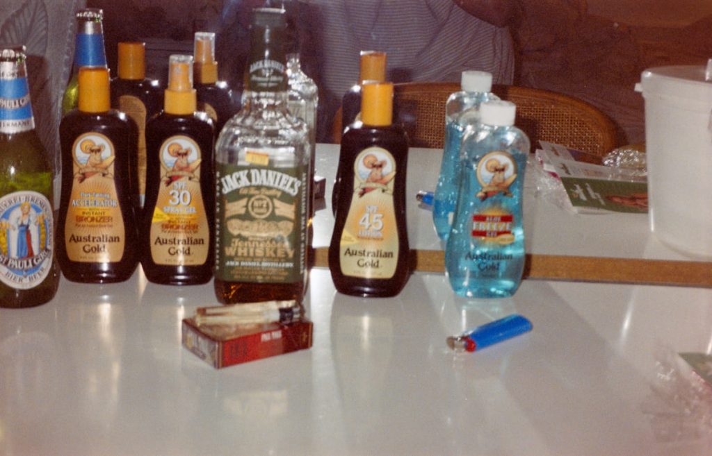 Flaschen mit Whiskey, Bier und Sonnencreme sowie Feuerzeuge auf einem weißen Tisch.