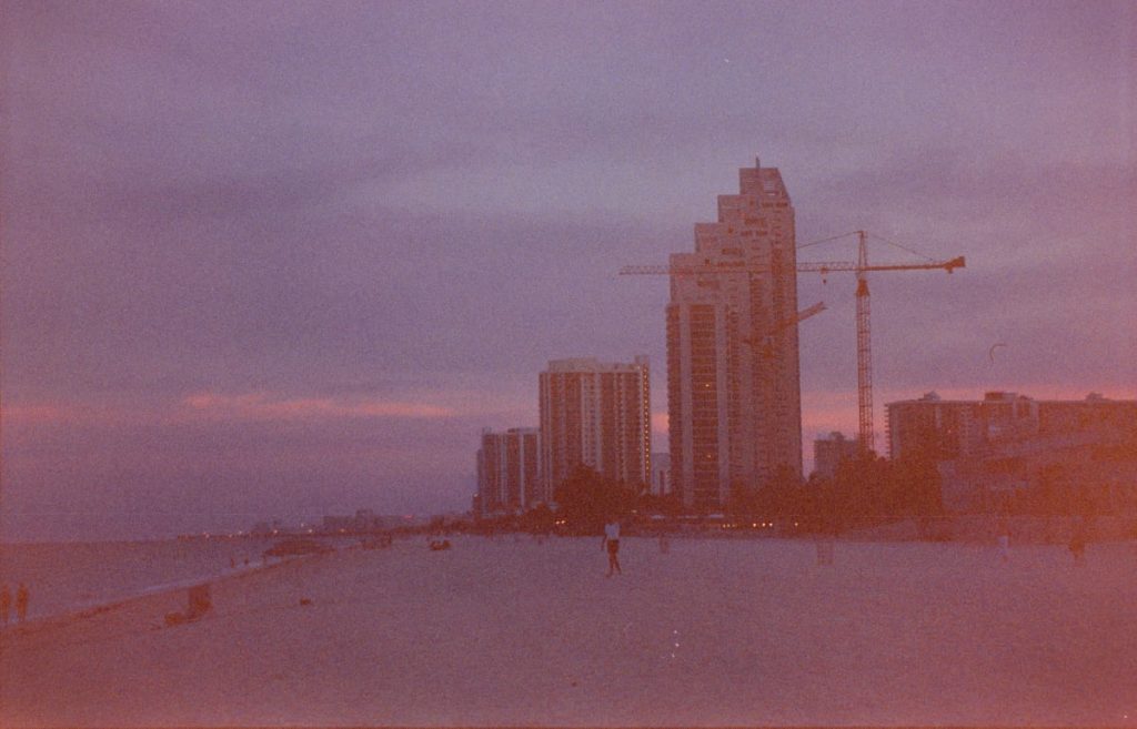 Weiter Strand in der Dämmerung mit Skyline-Gebäuden und einem Baukran im Hintergrund.