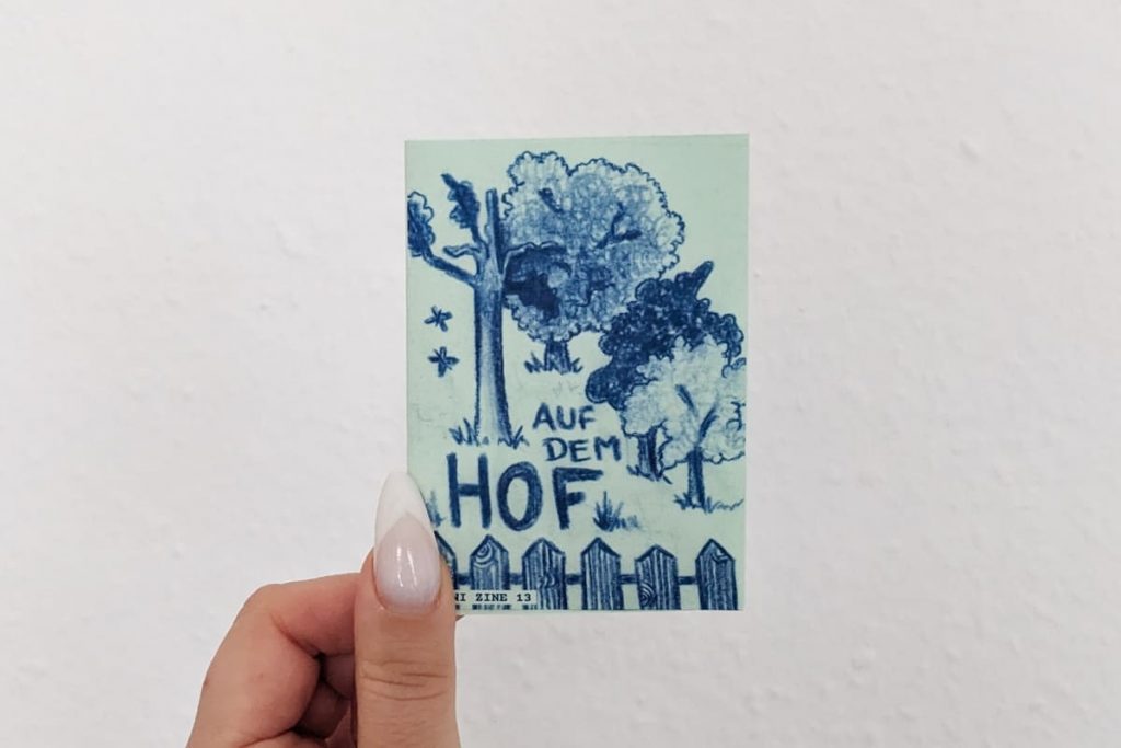 Hand hält Zine. Auf dem Titel: blaue Zeichnung mit Gartenzaun und vier Bäumen. Schriftzug "Auf dem Hof"
