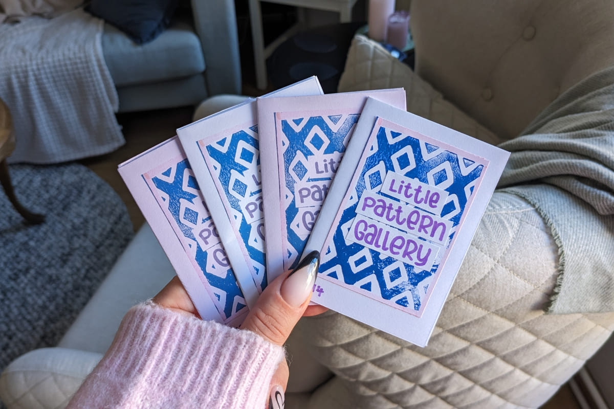 Hand mit rosa Ärmel hält vier Mini Zines. Auf dem Titel steht "Little Pattern Gallery", dahinter ein blauer Linoldruck mir Rautenmuster.