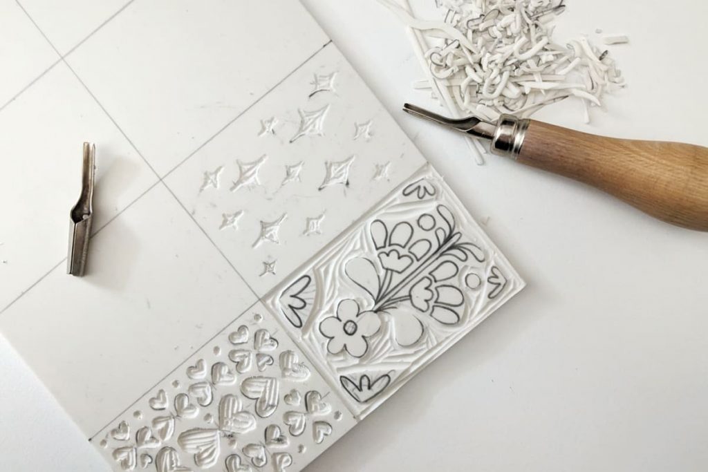 Linolschnittplatte und Werkzeug liegen auf dem Tisch. Auf der Platte sind erste Muster vorgezeichnet und ausgeschnitten. (Sterne, Herzen, Blume)