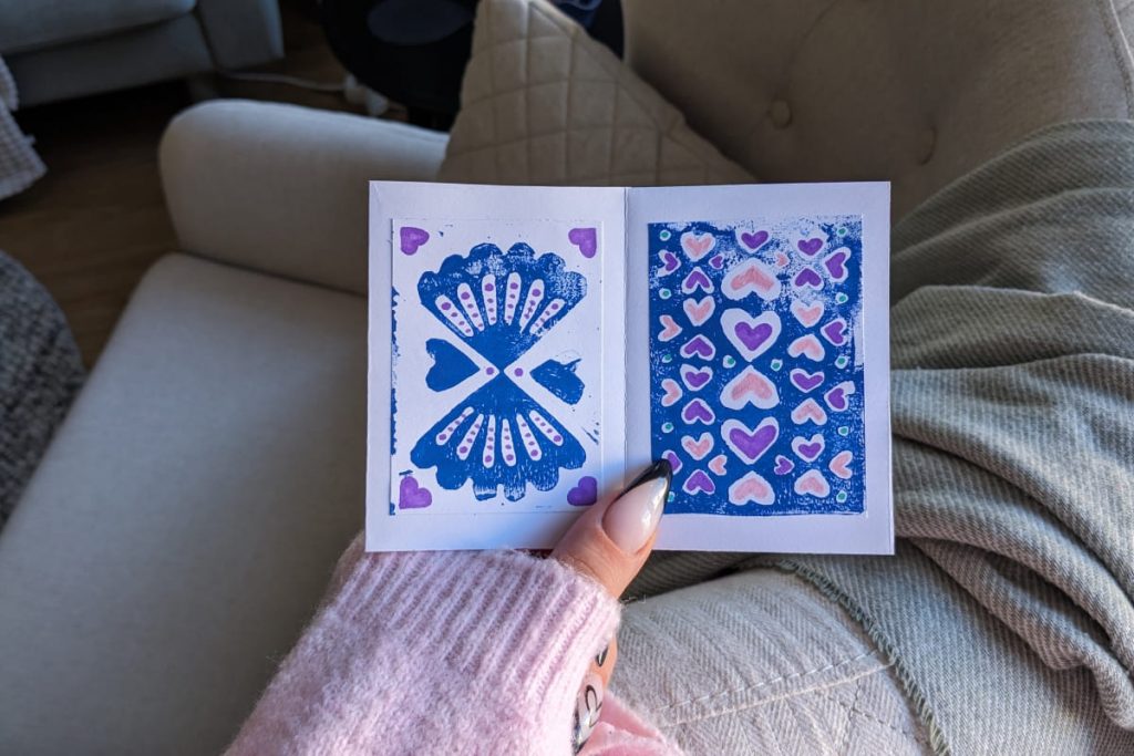 Hand mit rosa Ärmel hält offenes Zine. Zu sehen sind zwei blau gedruckte Linolschnitt-Muster, links Muscheln, rechts Herzen. Dekoriert und ausgemalt mit lila und rosa Filzstift.