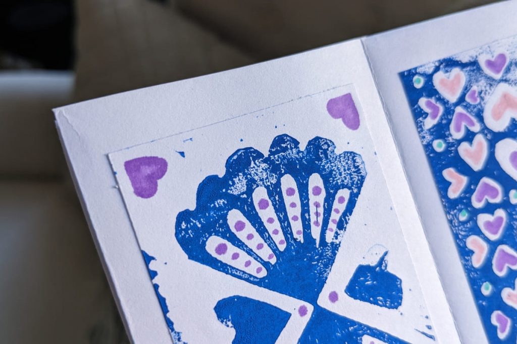 Nahaufnahme blau gedrucktes Linolschnitt-Muster mit Muscheln und dazugemalten lila Punkten und Herzen.