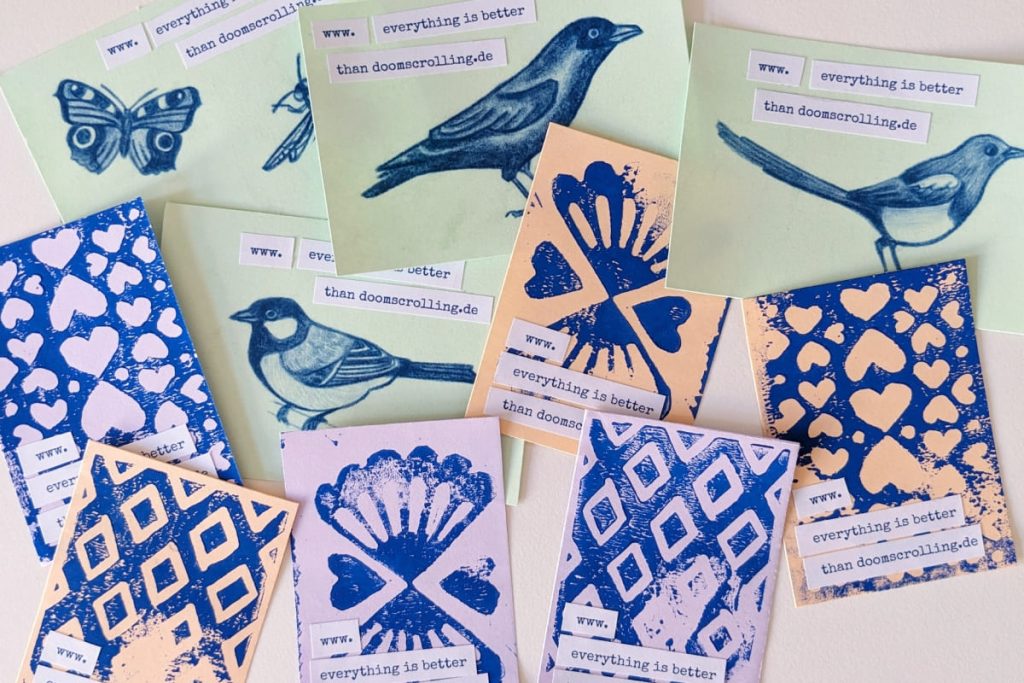 Visitenkarten aus blauen Linolschnitt-Musterdrucken auf lila und orangem Papier und Vogelzeichnungen auf grünem Papier. Dazu als Streifen aufgeklebt die Blog-URL.