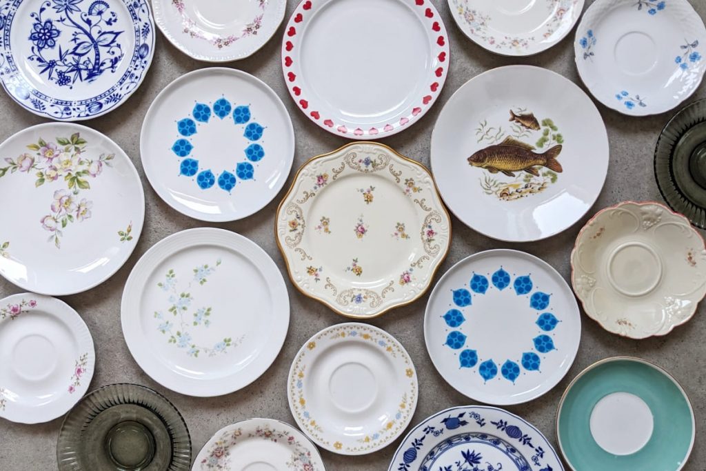 Vielfältige Sammlung dekorativer Porzellanteller, die auf einem grauen Boden. Verschiedene Designs, darunter blaue Zwiebelmuster, florale Motive in Rosa und Gelb, ein detailliertes Karpfen-Motiv, ein Teller mit einem roten Herzrand sowie modernere Muster mit blauen Tropfenformen.