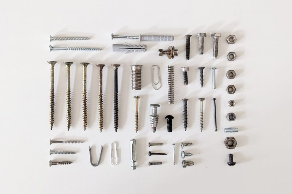 Rasterförmige sortierte Anordnung verschiedener Metall-Kleinteile auf weißem Hintergrund. Diverse Schrauben in unterschiedlichen Längen, Sechskantmuttern, zwei Büroklammern, eine Metallfeder, Kunststoffdübel, eine U-Klammer und kleine Nägel.