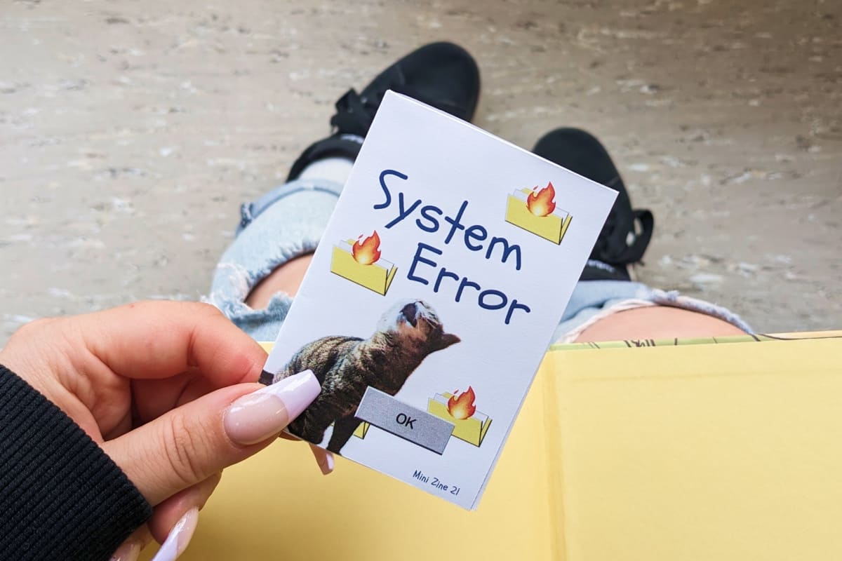 Person hält das Mini-Zine „System Error“ in der Hand. Cover: eine aufblickende Katze, brennende gelbe Aktenordner und eine klassische graue „OK“-Schaltfläche. Hintergrund: die Beine der Person in zerrissenen Jeans und schwarzen Sneakern.