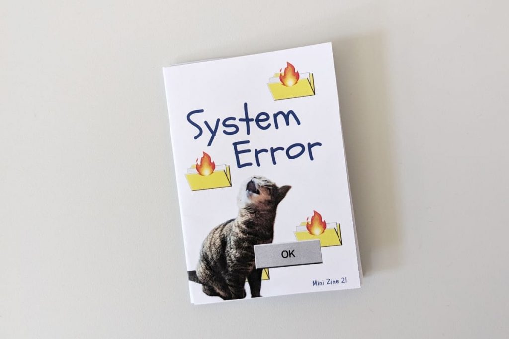 Zine-Cover „System Error“. Das Design erinnert an alte Computer-Betriebssysteme. Eine getigerte Katze schaut verzweifelt nach oben zu drei brennenden Ordner-Symbolen. Unten rechts steht klein „Mini Zine 21“.