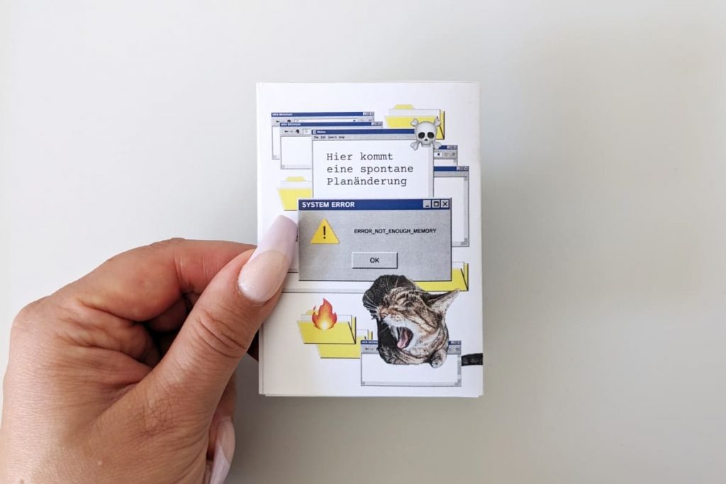 Rückseite: fauchende Katze vor einer Fehlermeldung mit dem Titel „SYSTEM ERROR“ und der Nachricht „ERROR_NOT_ENOUGH_MEMORY“. Darüber steht der Text: „Hier kommt eine spontane Planänderung“, ergänzt durch ein Totenkopf-Emoji.