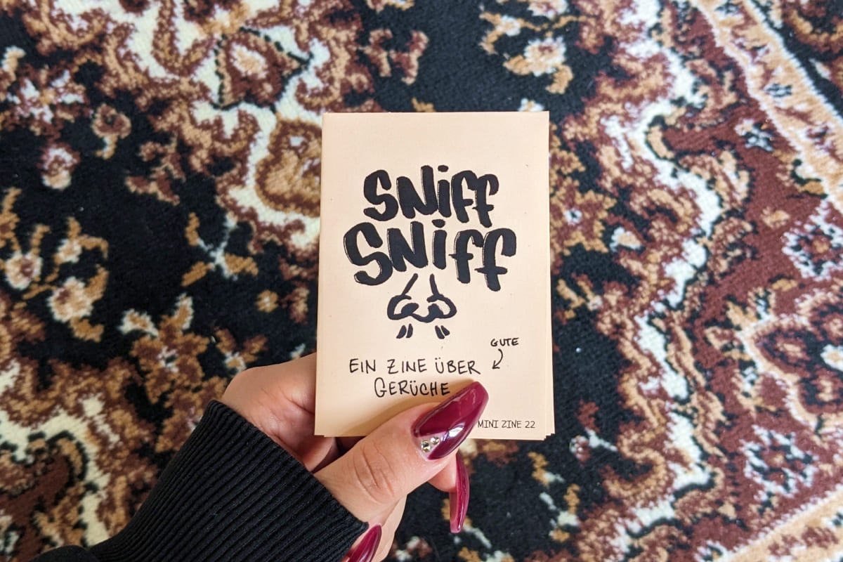 Hand hält Mini-Zine vor braunem Teppich mit Persermuster. Titel: „SNIFF SNIFF – EIN ZINE ÜBER GUTE GERÜCHE“ mit einer gezeichneten Nase.