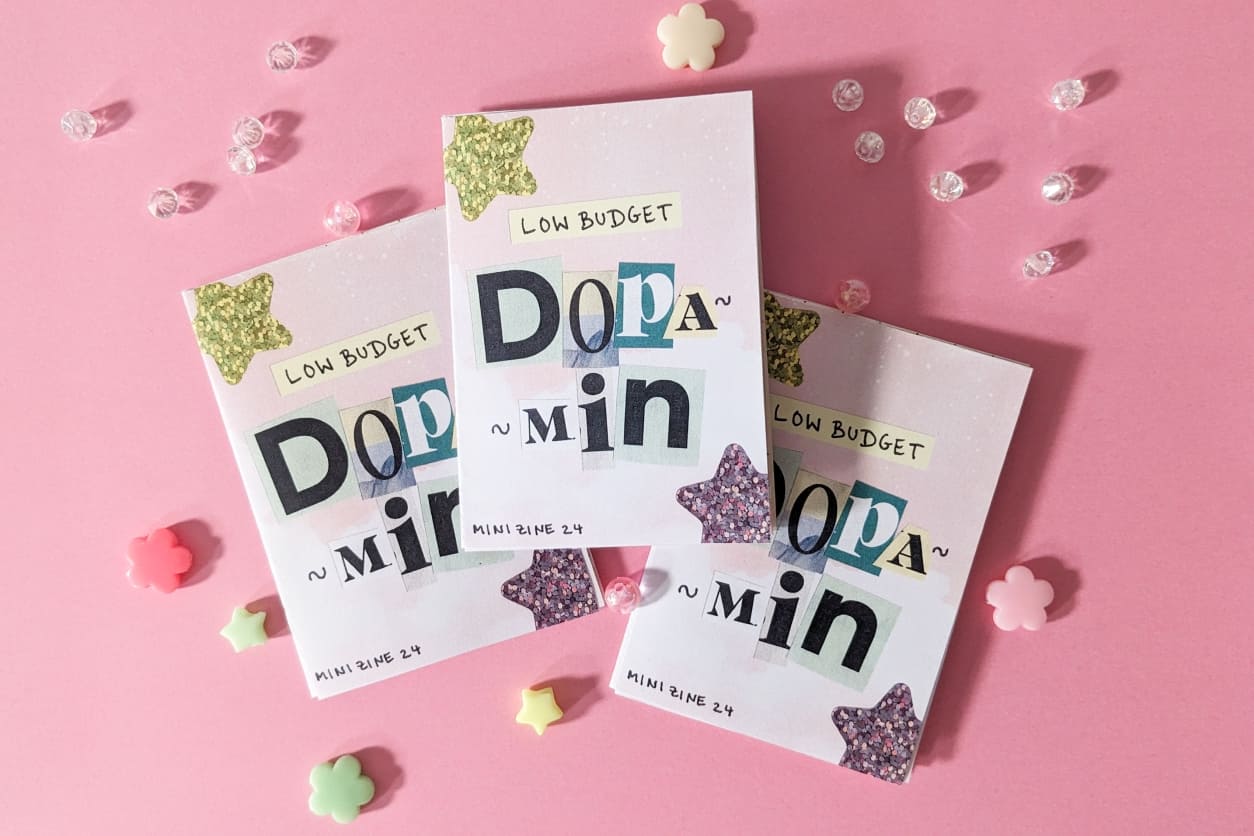 Drei pastellfarbene Mini-Zines mit ausgeschnittenen Buchstaben „Low Budget Dopamin“ und Glitzersternen, dekoriert mit Perlen auf pinkem Grund.