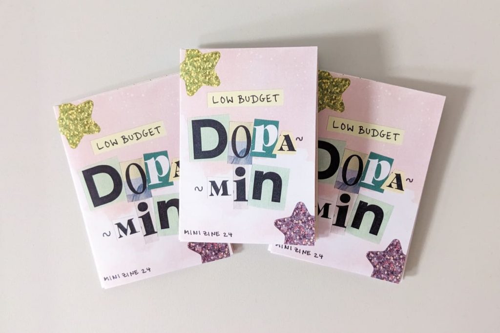 Drei Exemplare des Mini-Zines „Low Budget Dopamin“ mit Collagen-Schriftzug und Glitzersternen auf dem Titel.
