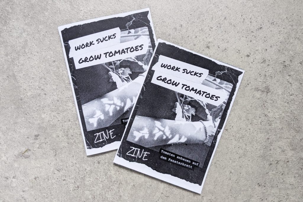 Zwei schwarz-weiße Zines mit kopiertem Grunge-Look und der Aufschrift „Work sucks, grow tomatoes“ liegen auf rauem Betonboden.