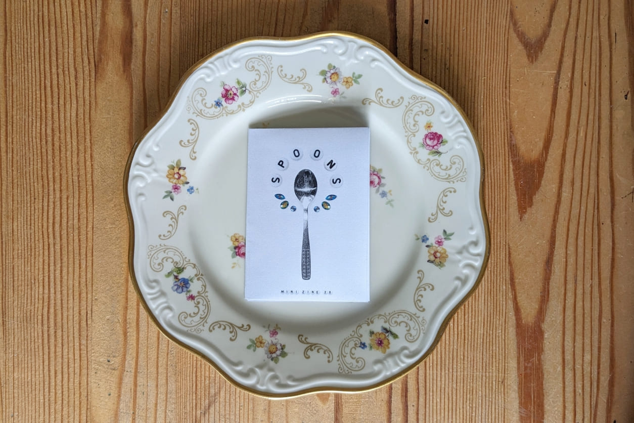 Weißes Mini-Zine mit Löffel-Abbildung liegt mittig auf einem blütenverzierten Teller mit Goldrand auf Holz.