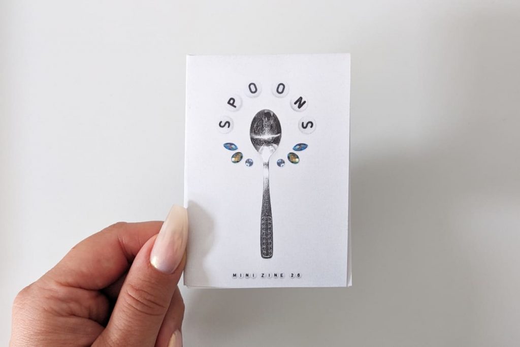 Eine Hand hält das Mini-Zine. Auf dem Cover: mittig ein freigestellter Löffel, drumherum im Halbkreis "SPOONS" aus Buchstabenperlen, unterer Halbkreis mit Schmucksteinen
