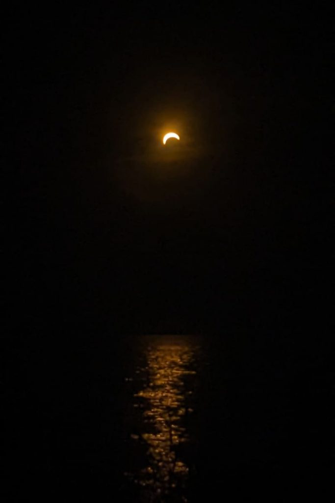 Schwarzes Bild mit sichelförmiger Sonne am Himmel, die vom Mond verdeckt wird. Im unteren Teil Spiegelungen im Wasser.