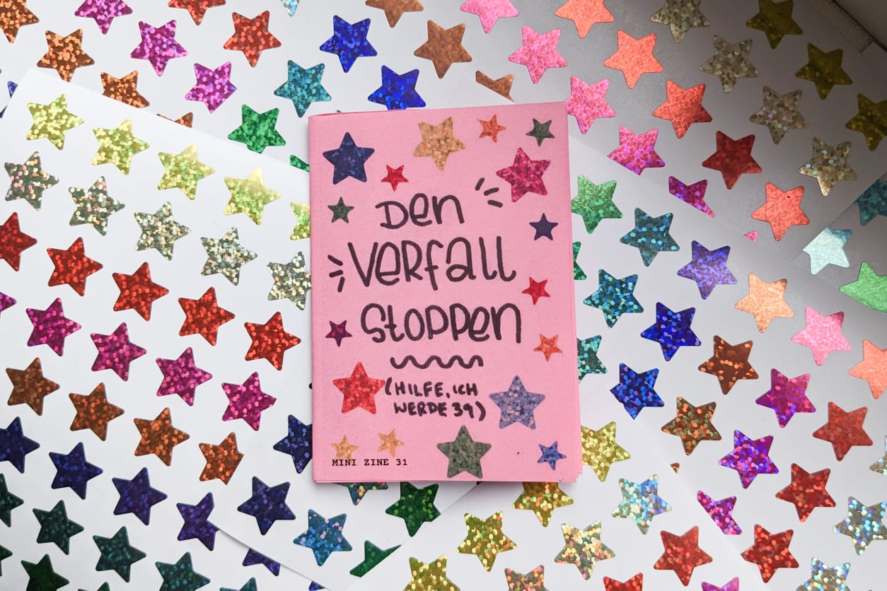 Ein pinkes Mini Zine liegt auf einem Untergrund aus unendlich vielen Glitzer-Sterne-Stickern