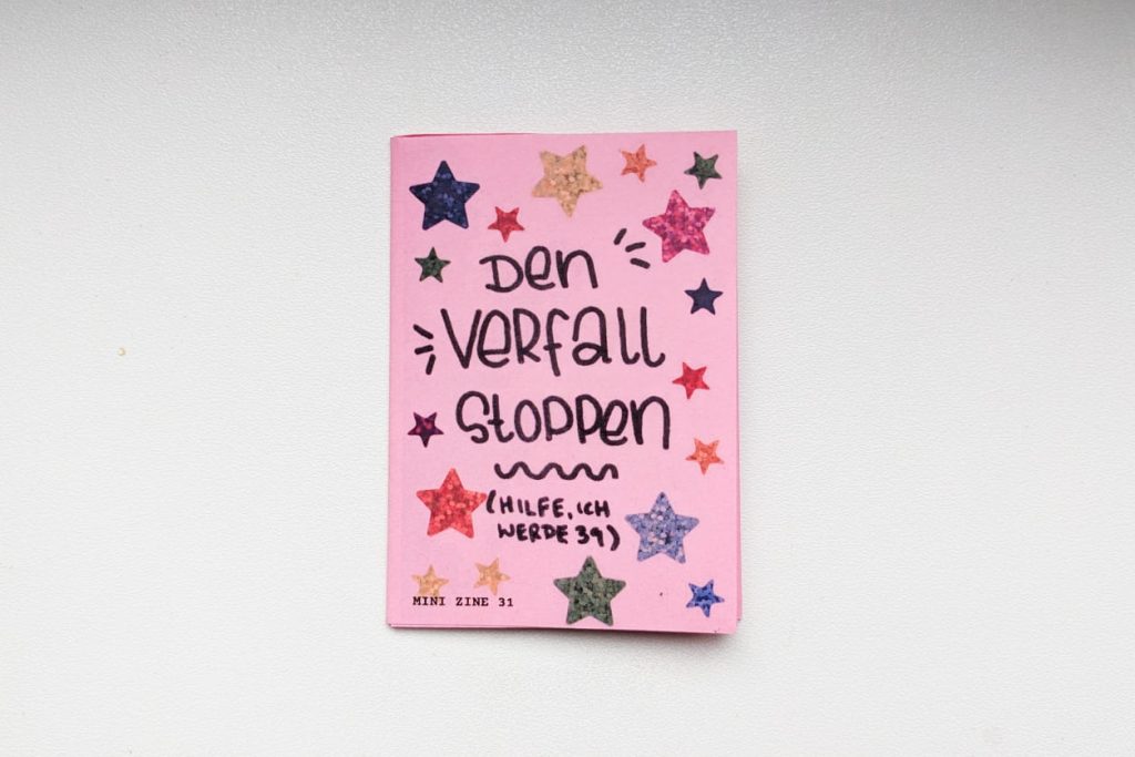 Pinkes Mini Zine. Titel handschriftlich: "Den Verfall stoppen (Hilfe ich werde 39)", dekoriert mit bunten Glitzer-Sterne-Stickern.