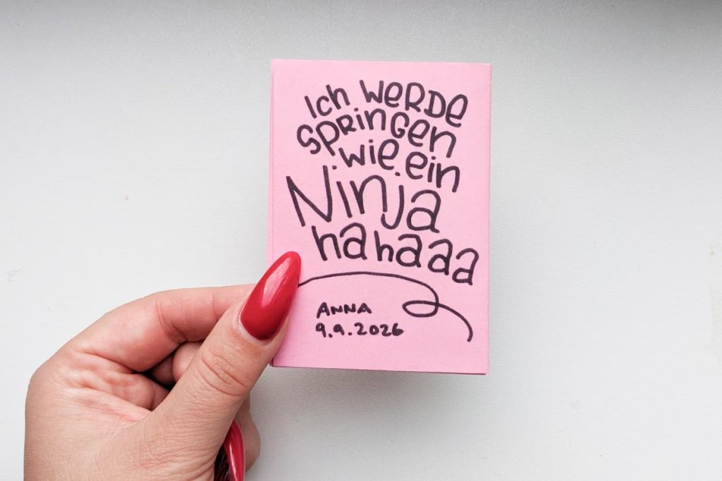 Hand mit roten langen Nägeln hält das Mini Zine. Rückseite handgeschrieben: Ich werde springen wie ein Ninja hahaaa. Anna 9.9.2026