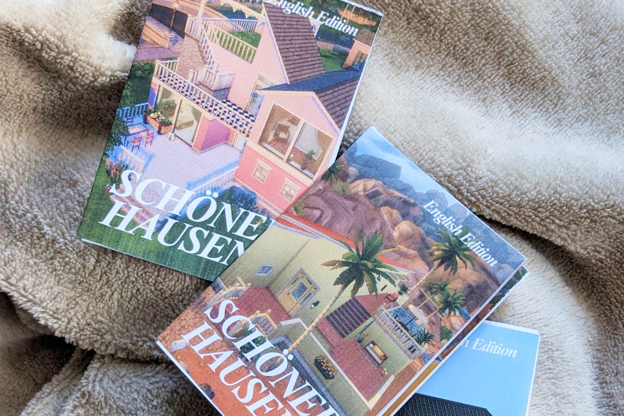 Drei englischsprachige Zines "Schöner Hausen" mit Sims-Häusern auf den Covern liegen auf einer beige-braunen Decke