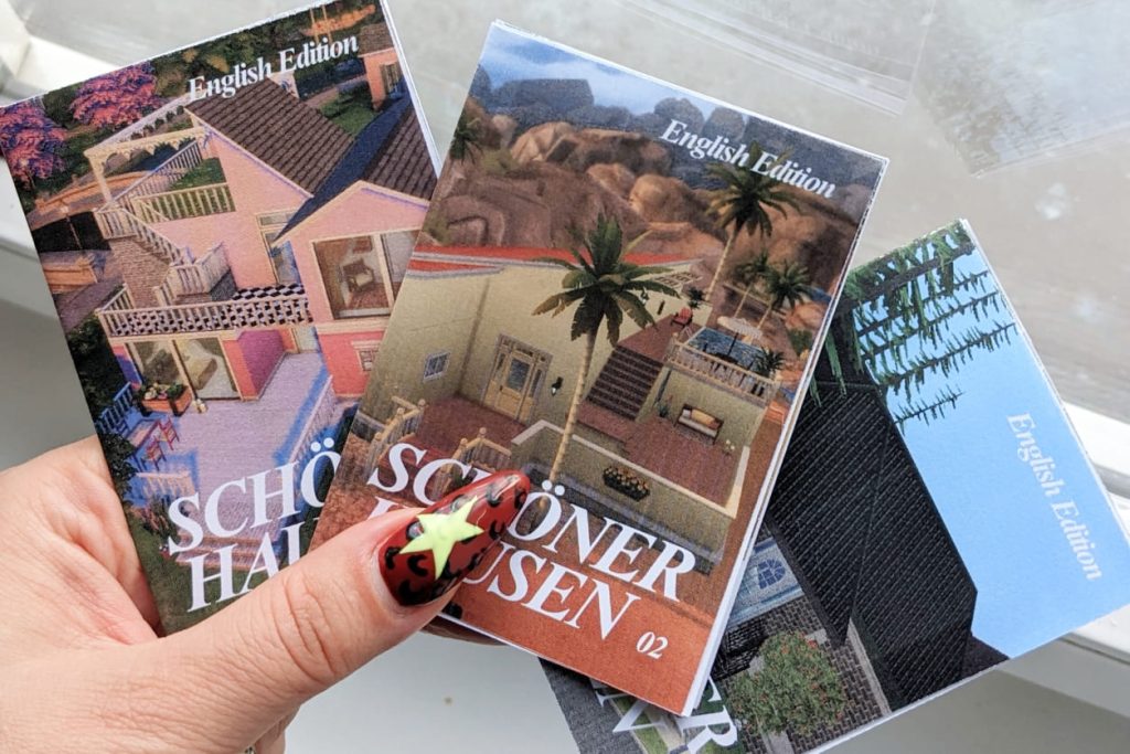 Hand mit auffälligem braunem Leo-Nagellack und neongelben Sternen hält drei kleine Zines mit Sims-Häusern und dem Titel 'SCHÖNER HAUSEN'.