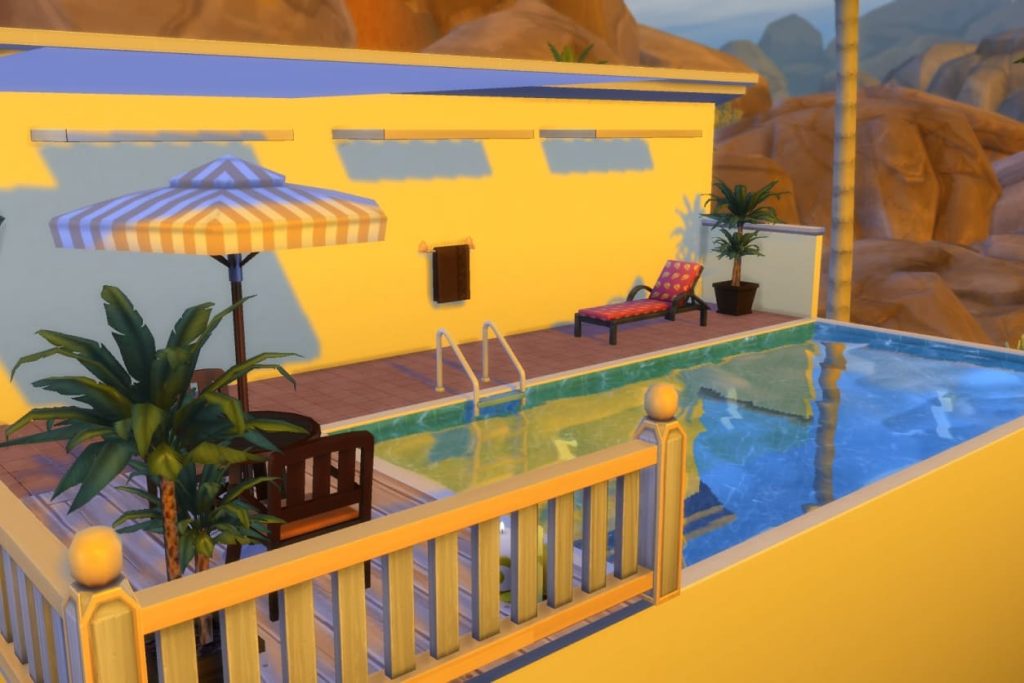 Blick auf eine Pool-Terrasse mit einem blauen Pool, Sonnenliege, Topf-Palmen, einem Esstisch mit gelb-weiß gestreiftem Sonnenschirm und einer felsigen Landschaft im Hintergrund.